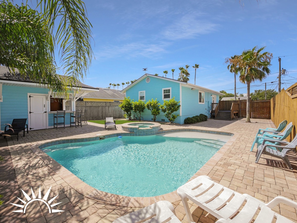 A private pool area is surrounded by a sunlit patio, featuring lounge chairs for relaxation. The aqua pool is complemented by a raised hot tub nearby. Lush greenery and palm trees create a tropical atmosphere, while a charming blue house is visible in the background.