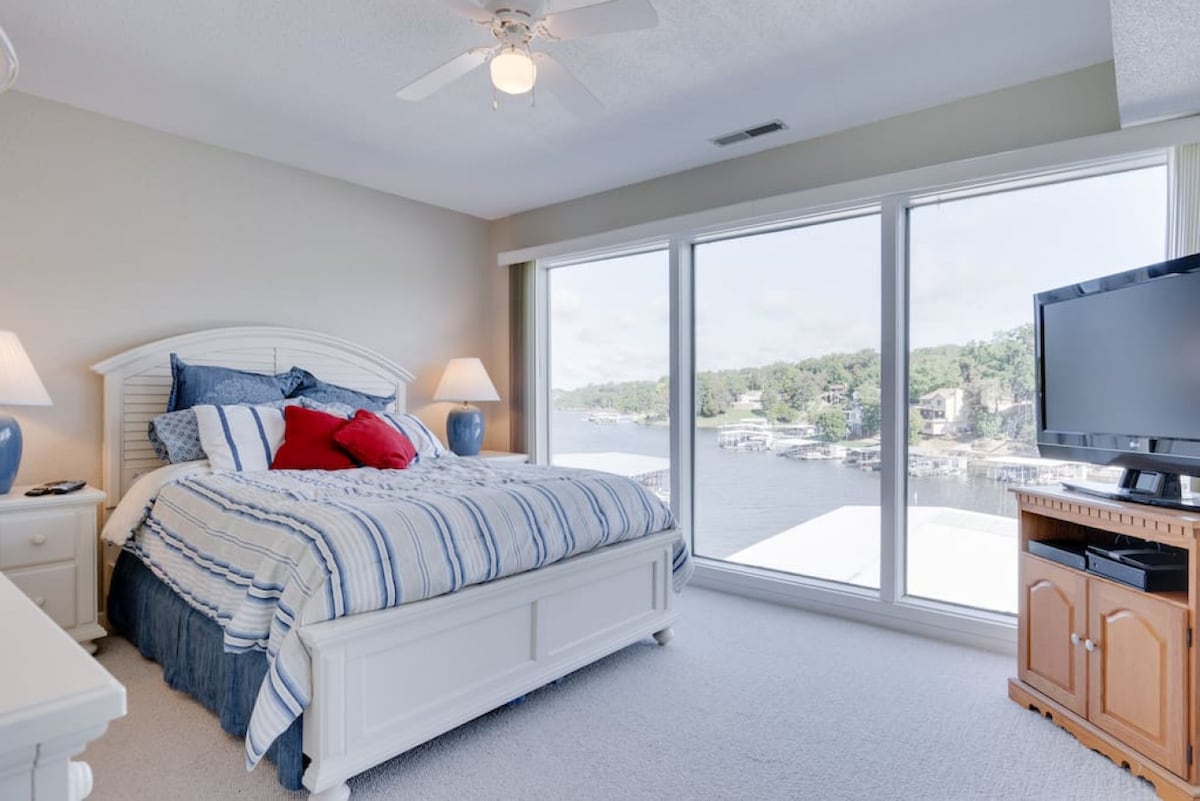A spacious bedroom features a queen bed with blue and white striped bedding. Large windows provide views of the water, allowing natural light to flood the room. A flat-screen TV is positioned on a wooden stand, with bedside lamps adding a soft glow.