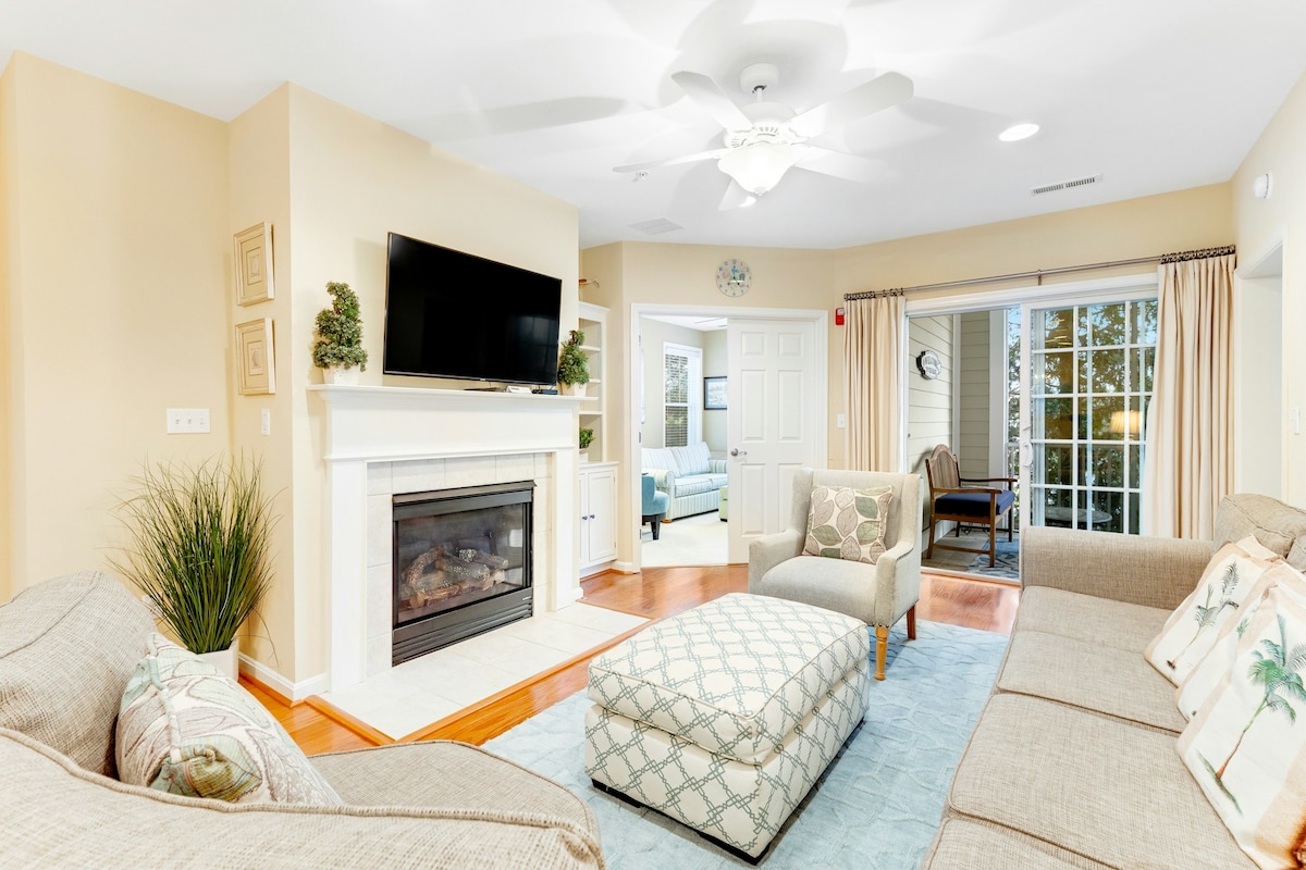 A spacious living area features a light-colored sectional sofa and an upholstered ottoman. A flat-screen television is mounted above a cozy fireplace. Natural light enters from glass doors leading to a screened balcony, enhancing the room's inviting atmosphere.