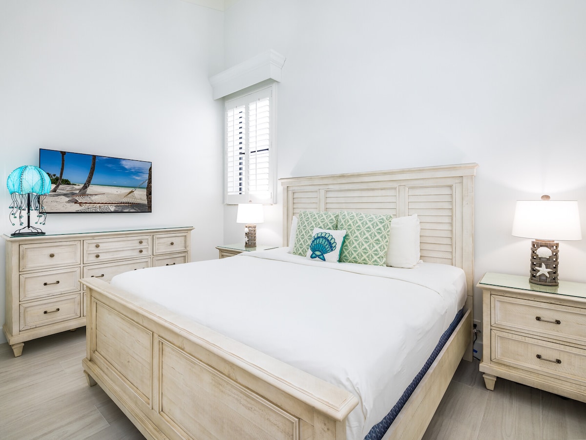 A well-appointed room features a spacious bed with white linens positioned against a bright wall. On either side of the bed, bedside tables with lamps add illumination. A dresser complements the decor, while a TV mounted above adds entertainment options.