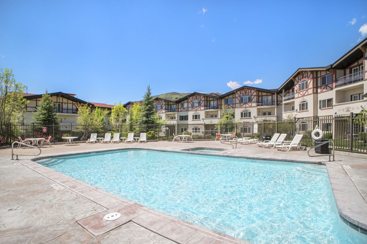 An outdoor swimming pool is surrounded by a spacious deck with lounge chairs and greenery. The villa's building is visible in the background, showcasing a blend of architectural styles and landscaping under a clear blue sky.