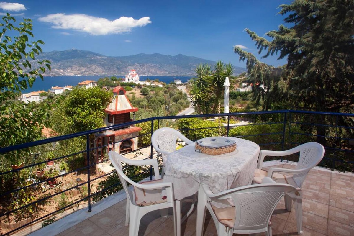A scenic balcony view features a round table surrounded by four chairs, overlooking lush greenery and a panoramic vista of the sea and distant mountains. A stone fireplace is positioned nearby, complementing the tranquil outdoor setting.