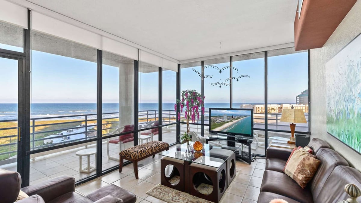 A spacious living room is framed by large glass windows, offering panoramic views of the ocean. Comfortable seating is arranged around a modern coffee table, with a flat-screen TV positioned for easy viewing. Natural light floods the space, enhancing the relaxed ambiance.