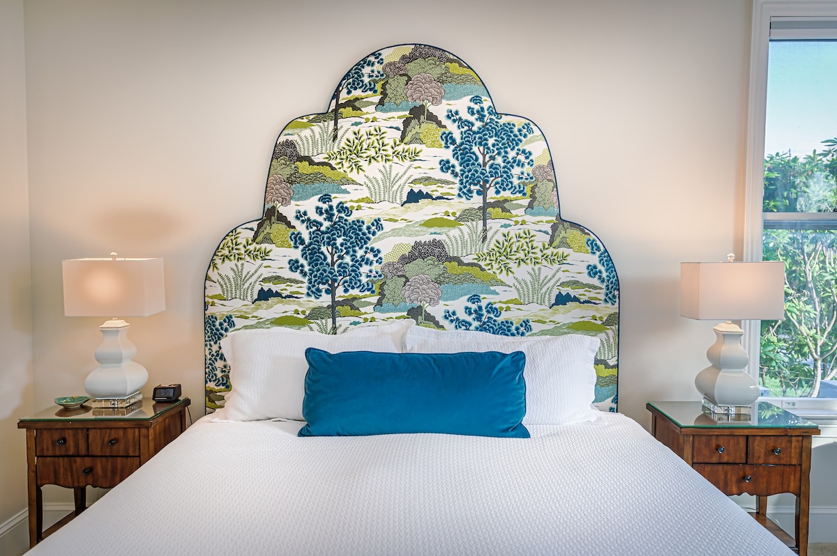 A spacious bed is framed by a decorative headboard featuring a floral pattern in blue and green. Two bedside tables with lamps are placed on either side, illuminating the serene sleeping area. A large window is visible, allowing natural light to fill the room.
