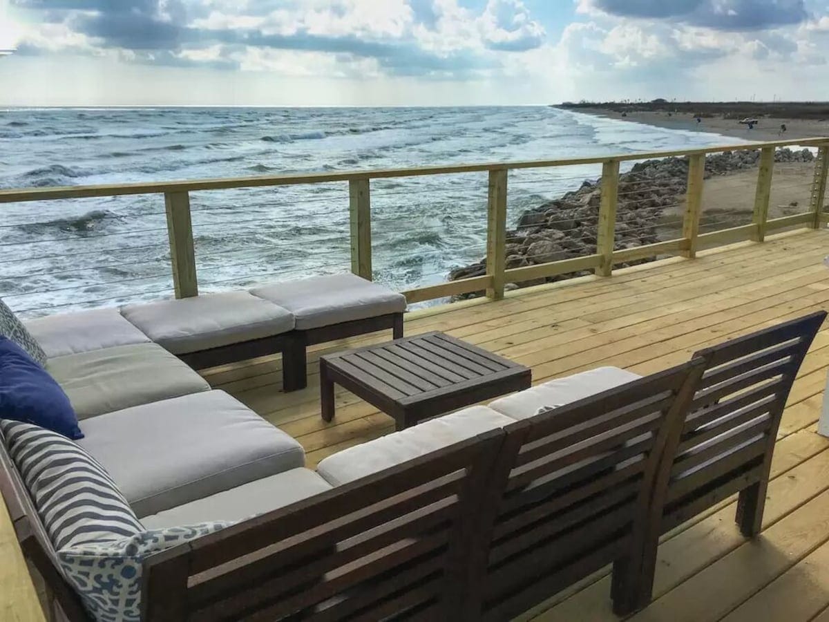 A spacious outdoor seating area is set on a wooden deck, overlooking the ocean. The seating arrangement includes a sectional couch with soft cushions and a small table placed at the center. The expansive view showcases gentle waves and a clear sky dotted with clouds.