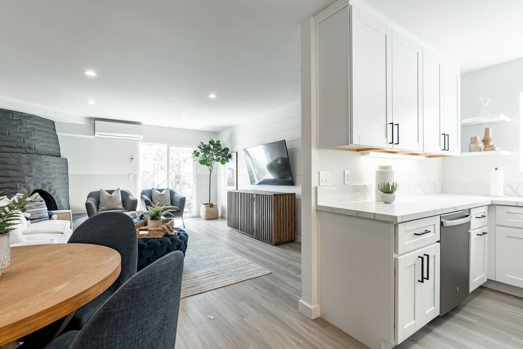 An open living space features a modern kitchen with white cabinetry and a marble countertop. The dining area includes a round table surrounded by dark chairs. A cozy seating area has a large couch, a sleek entertainment center, and large windows allowing natural light.