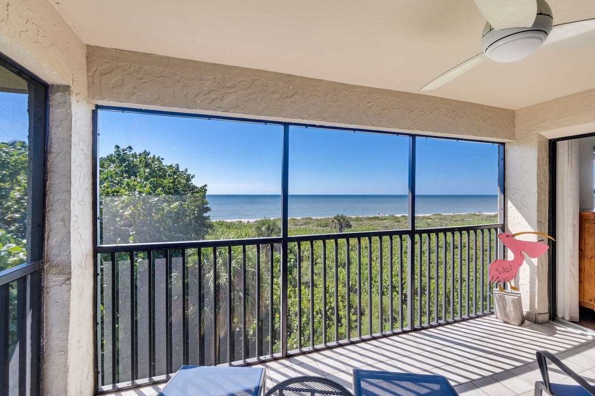 A screened-in lanai features a railing with expansive views of the Gulf waters and surrounding tropical foliage. Sunlight brightens the space, highlighting a small table and chairs designed for relaxation while enjoying the coastal scenery.