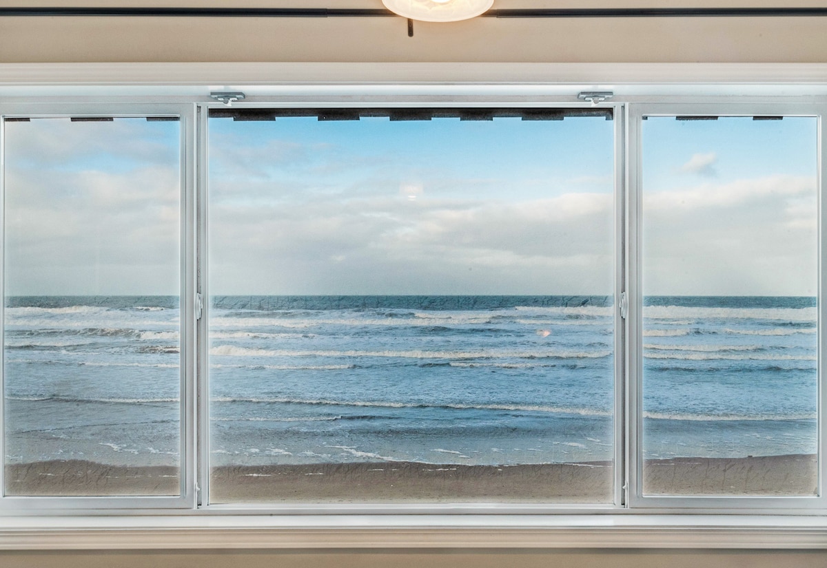 A panoramic view of the ocean is visible through large multi-pane windows. The scene showcases rolling waves and a sandy beach, with cloudy skies overhead. The tranquility of the ocean is complemented by the inviting light filling the room.