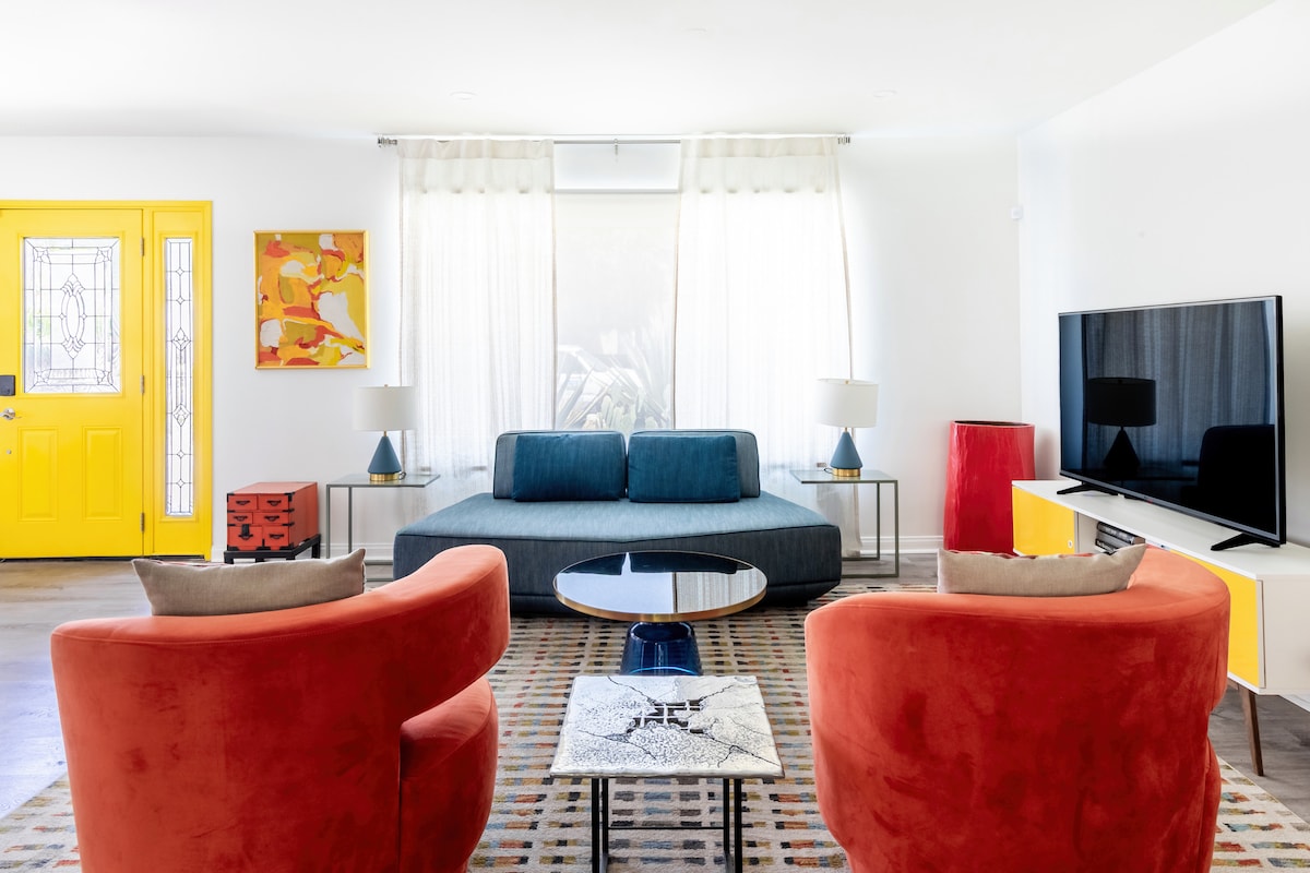 A bright living area features a low, spacious seating arrangement in blue and bold red accent chairs. Natural light streams through large windows, highlighting vibrant artwork on the walls. A sleek television is mounted, accompanied by a contemporary media console and an eclectic coffee table.