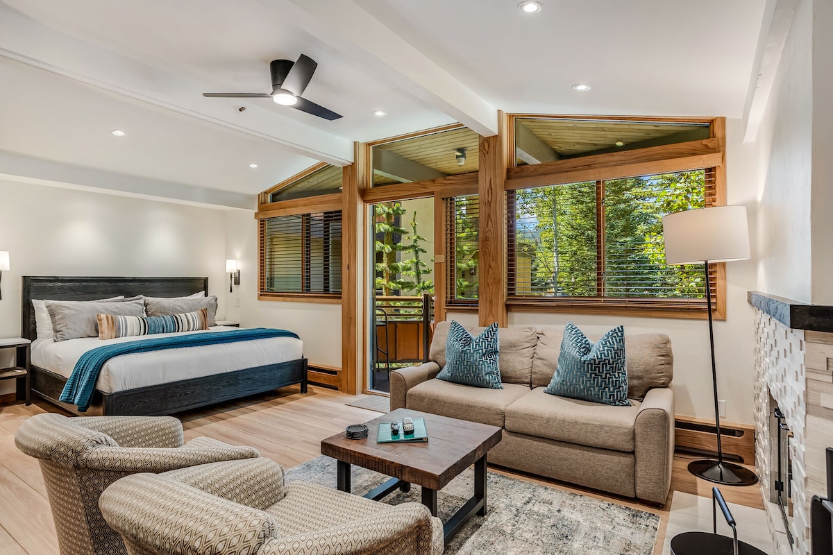 A spacious studio features large windows that bring in natural light and offer views of the greenery outside. The sleeping area includes a king bed with accent pillows, while a cozy seating area displays two armchairs and a sofa. A modern fireplace complements the inviting atmosphere.