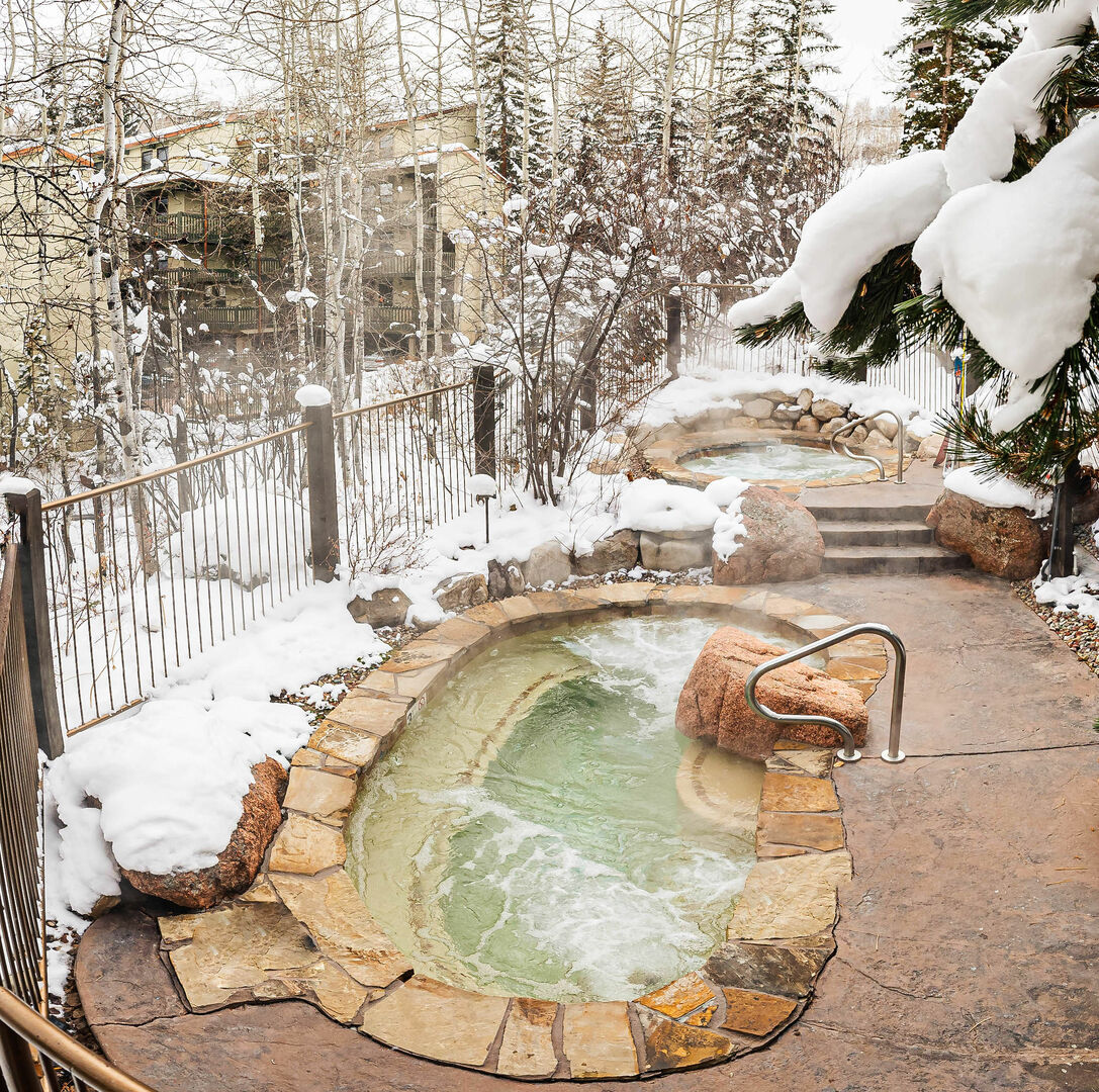 The outdoor hot tubs are surrounded by a snow-covered landscape and trees, featuring natural stone edges. Gentle steam rises from the water, creating a serene atmosphere, while the pathway leading to the tubs is lined with snow-dusted rocks and foliage.