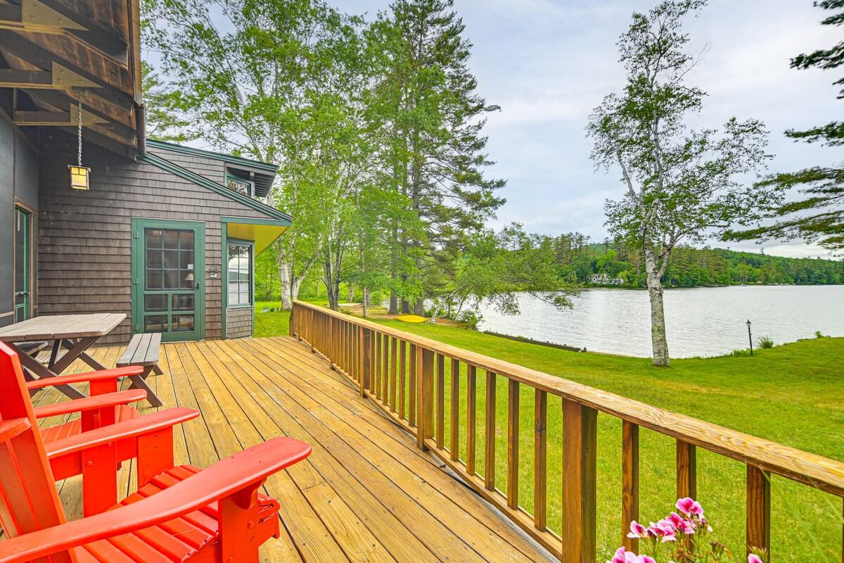 A spacious wooden deck is shown, featuring bright red Adirondack chairs overlooking a serene waterfront. Lush green grass and trees are visible in the background, alongside a calm body of water, creating a relaxing outdoor setting.