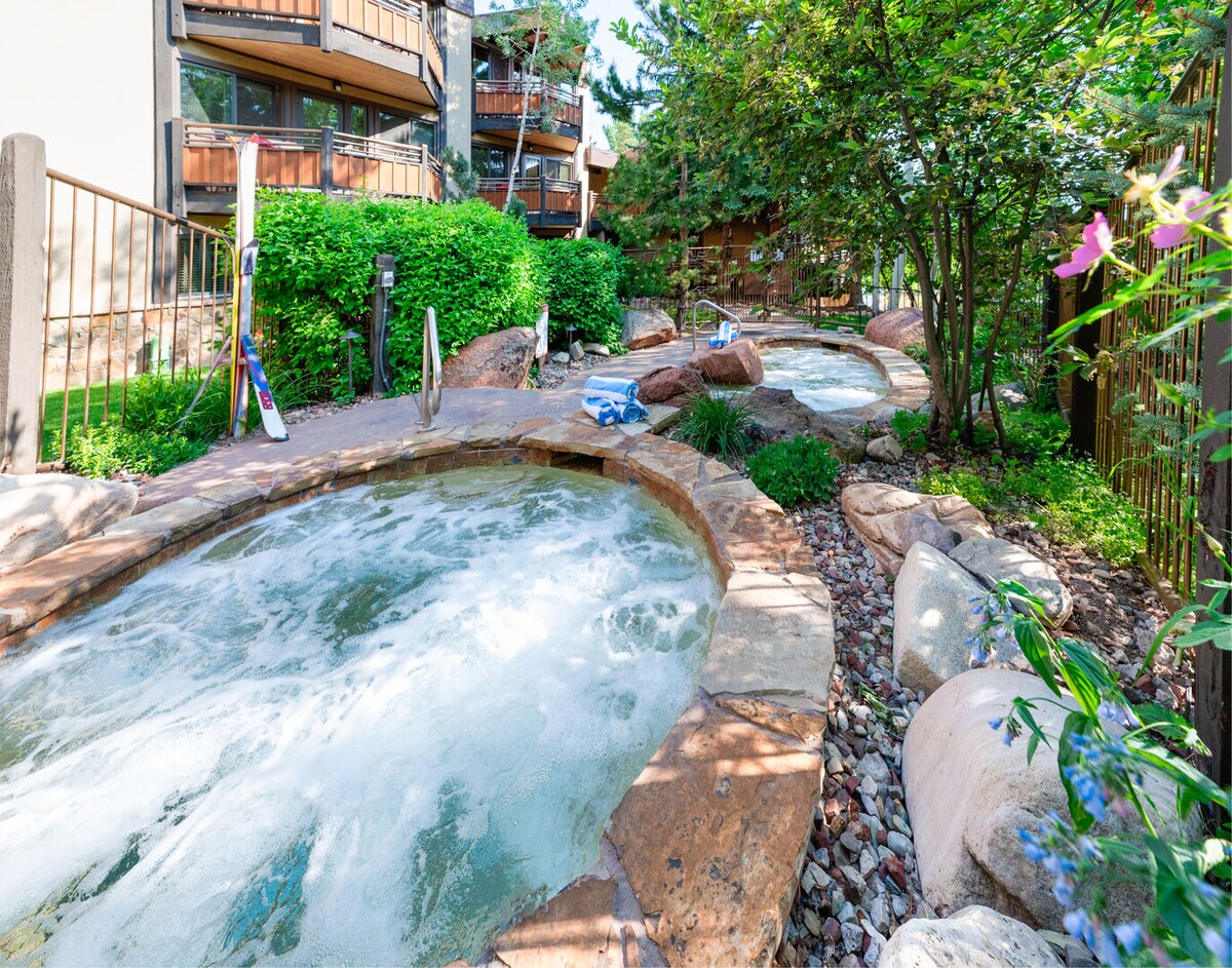 Two-tiered hot tubs are surrounded by lush greenery and natural stone pathways. Water bubbles invitingly within the tubs, set against a backdrop of well-maintained landscaping featuring various rocks and plants, creating a relaxing outdoor atmosphere.