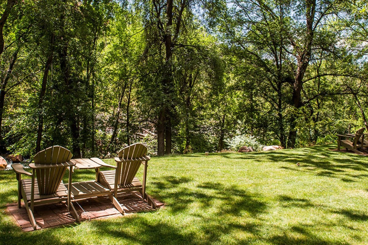 A serene outdoor space features two wooden lounge chairs positioned on a grassy area. Sunlight filters through the surrounding trees, casting dappled shadows on the lawn. The vibrant greenery enhances the natural setting, creating a tranquil retreat.