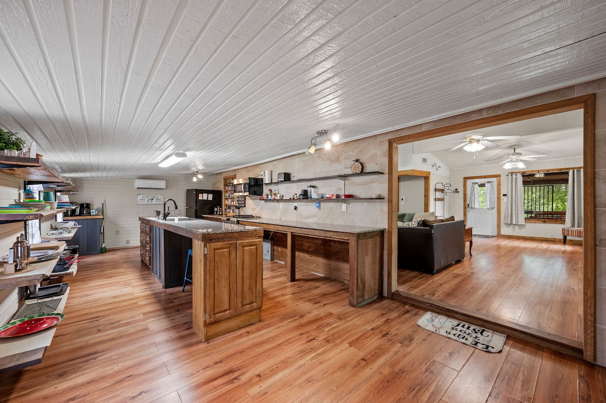 An open kitchen and living area feature warm wood flooring and white ceiling beams. The kitchen has a large island with a wooden countertop and ample shelving. The living space showcases comfortable seating, with natural light entering through a window overlooking the outdoors.