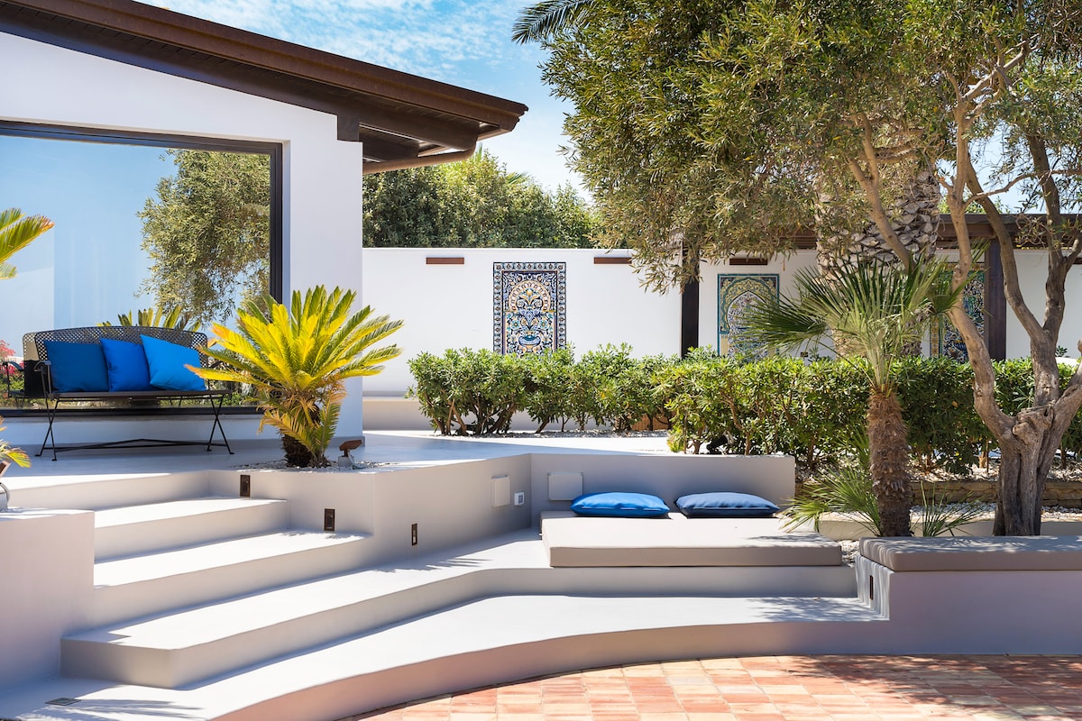 A spacious outdoor area is featured, showcasing stepping stones leading to various seating options. Lush greenery surrounds the area, and decorative tiles add character to the villa's façade. Bright blue cushions provide comfortable resting spots amidst the serene environment, with clear skies above.