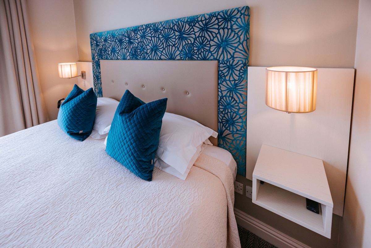 A cozy guest room features a spacious bed with a light-colored quilt and decorative blue accent pillows. The headboard displays a patterned design in shades of blue. Warm light is provided by two bedside lamps mounted on white nightstands.