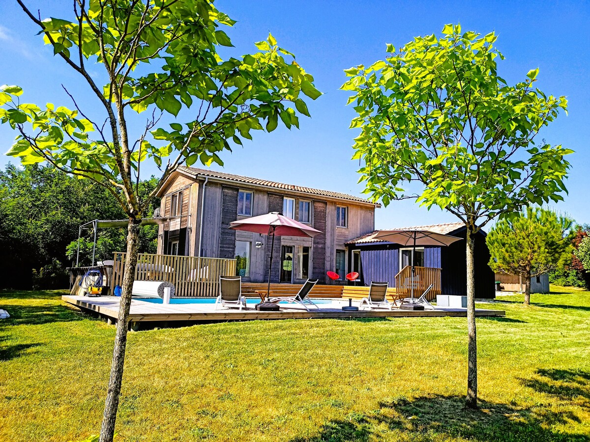 A contemporary wooden house sits surrounded by greenery, with a spacious deck featuring lounge chairs and an umbrella. Two small trees frame the scene, while a private heated pool is visible in the background, set against a clear blue sky.