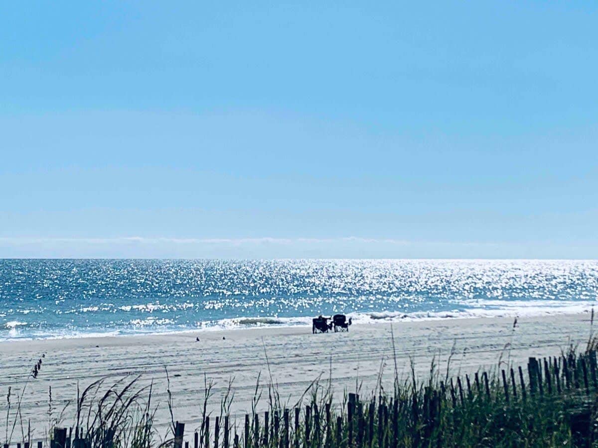 A serene beach scene is depicted with soft waves gently lapping at the shoreline. A few beach chairs are positioned near the water's edge, with the horizon reflecting sunlight. Beach grass gently sways in the foreground under a clear blue sky.