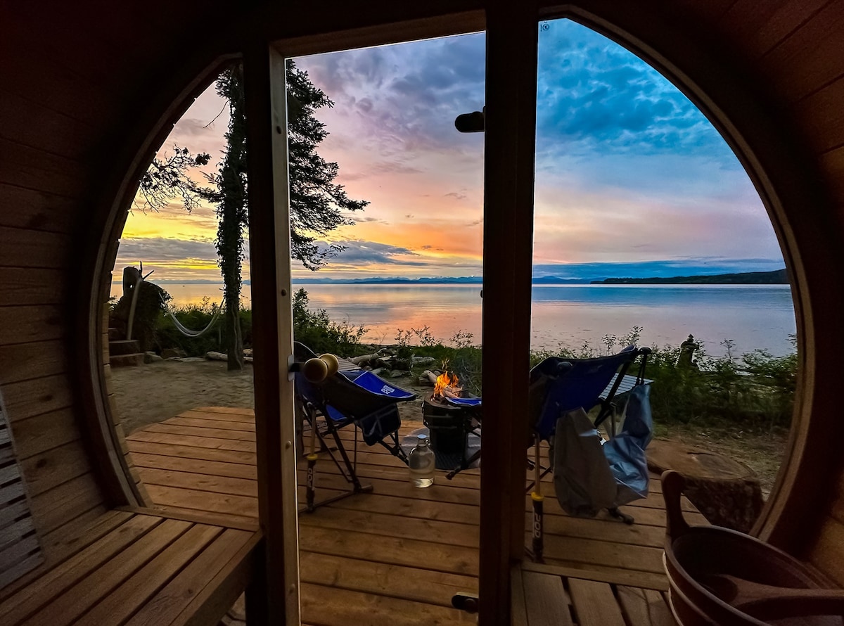 A circular wooden doorway frames a view of a serene lake at sunset, with hues of orange and purple reflected on the water. Two blue chairs are positioned around a fire pit on the deck, surrounded by lush greenery and the calming landscape.
