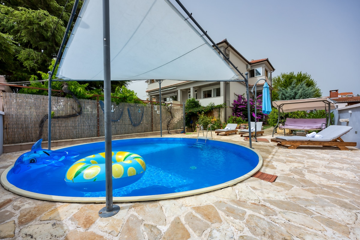 A circular pool is surrounded by a stone patio and shaded by a canopy. Sun loungers and a swing are positioned nearby, while a colorful pool float adds a playful element to the clear blue water.
