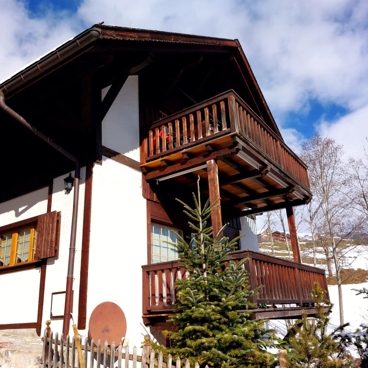 The exterior of the chalet features a combination of wood and white-washed walls, showcasing two balconies with wooden railings. Lush green trees are visible in the foreground, contrasting with the bright blue sky and white clouds above.