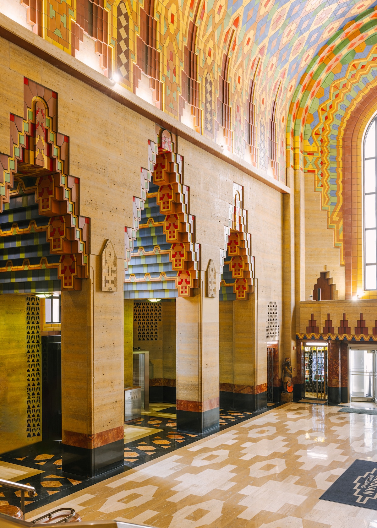 The historic lobby is adorned with vibrant Art Deco details, showcasing colorful geometric patterns on the walls and ceiling. Elegant archways frame the entrances, while polished marble floors reflect the intricate design, contributing to the unique architectural character of the space.