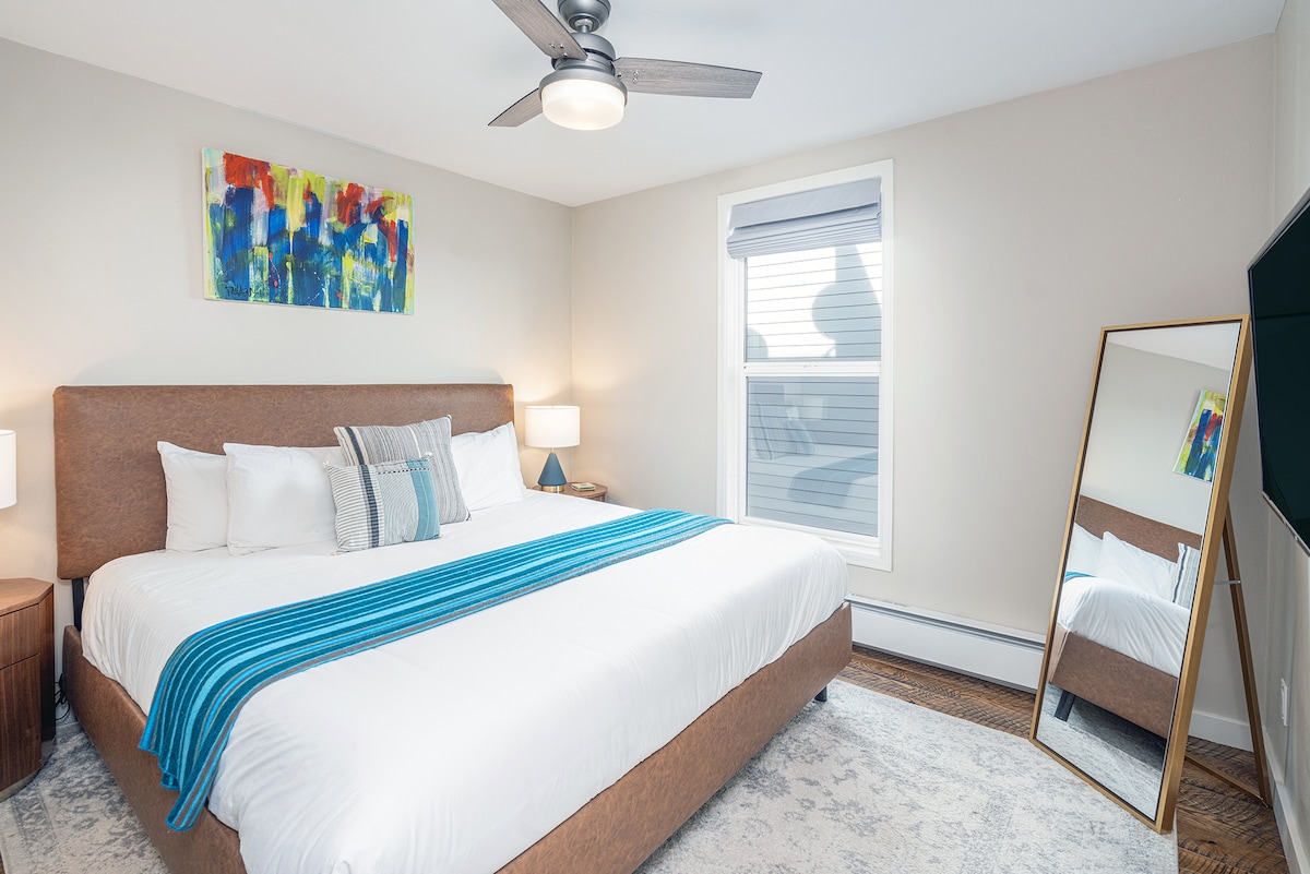 A well-lit bedroom features a spacious bed with crisp white linens, complemented by a blue accent blanket. A large mirror reflects the room's neutral tones, while colorful artwork adds a touch of vibrancy. Natural light filters through the window, enhancing the inviting ambiance.