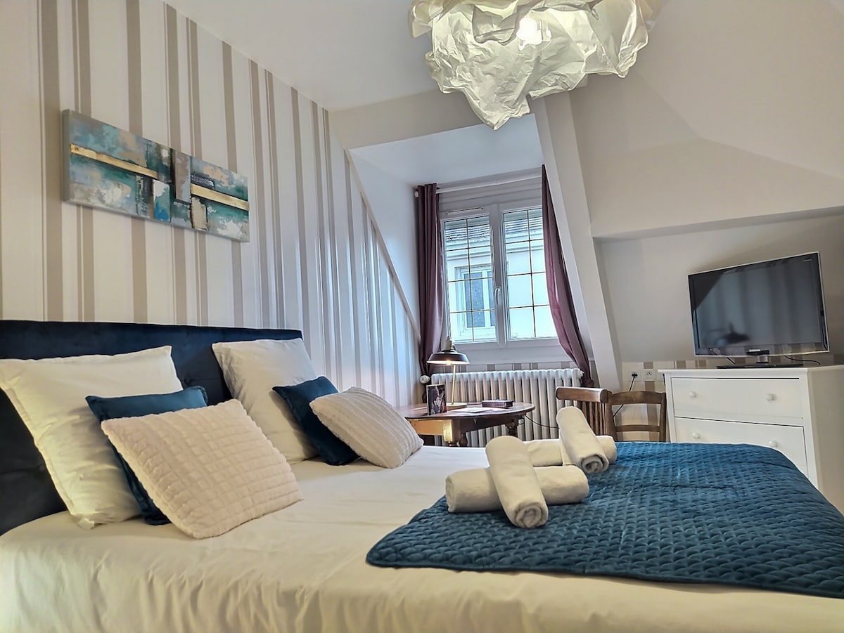A well-appointed bedroom is displayed, featuring a large bed with decorative pillows and a textured blanket. Soft lighting is provided by a unique ceiling fixture. A flat-screen TV and a small table with chairs are visible, along with windows allowing natural light into the space.