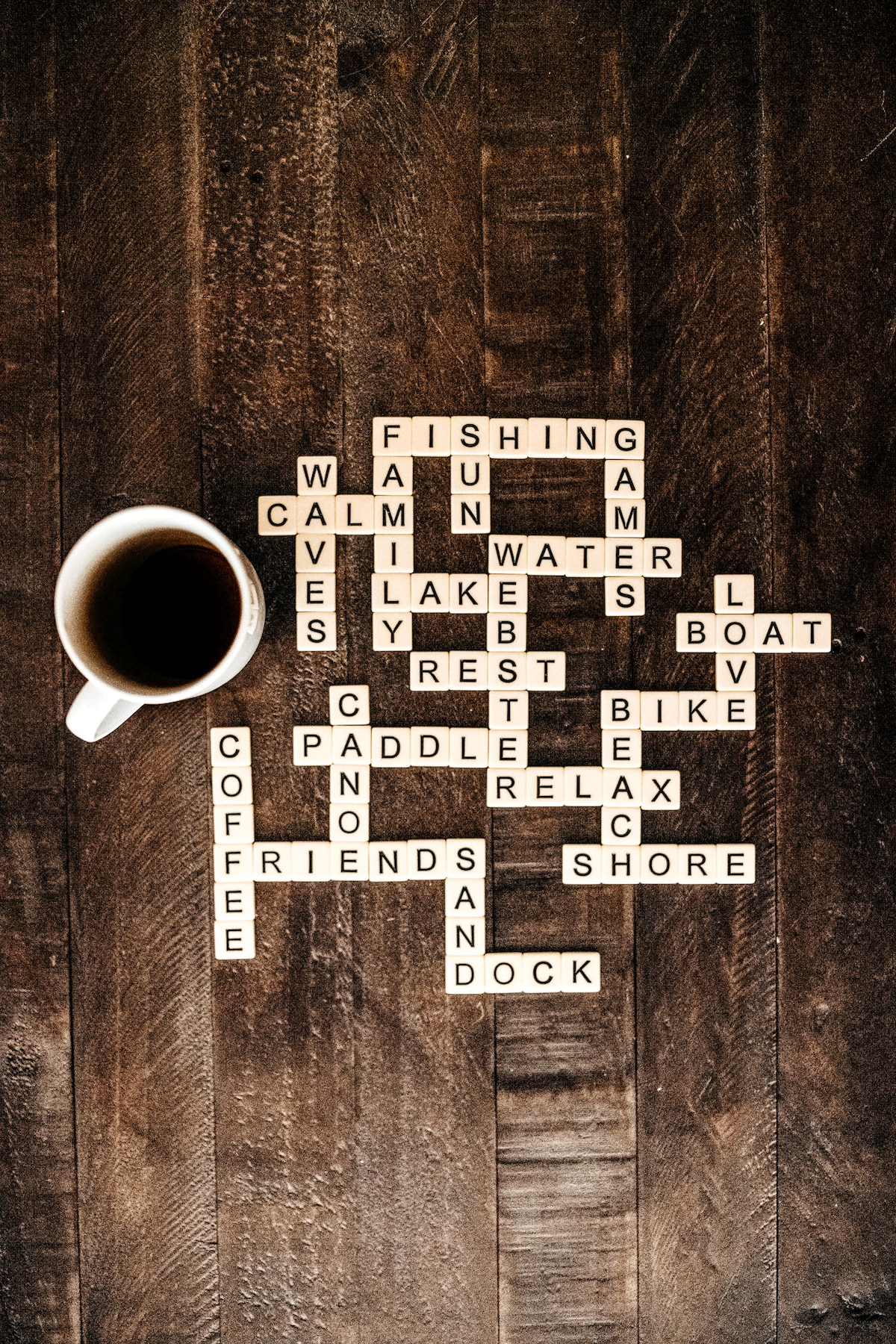 A coffee mug rests on a wooden table, surrounded by lettered tiles arranged to spell out various words related to leisure activities, such as 'fishing,' 'paddle,' and 'relax.' The rustic brown surface of the table adds to the relaxed atmosphere.
