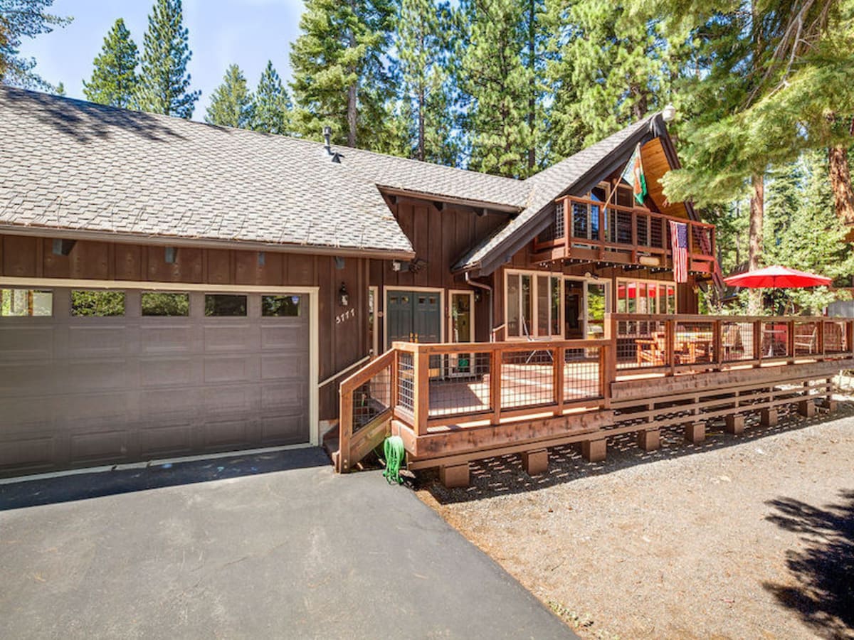 A spacious cabin exterior is surrounded by tall trees, highlighting the wooden structure and sloped roof. A large front deck features upgraded furnishings and an umbrella for shade. A well-maintained driveway leads to a garage door on the left, emphasizing the inviting outdoor space.