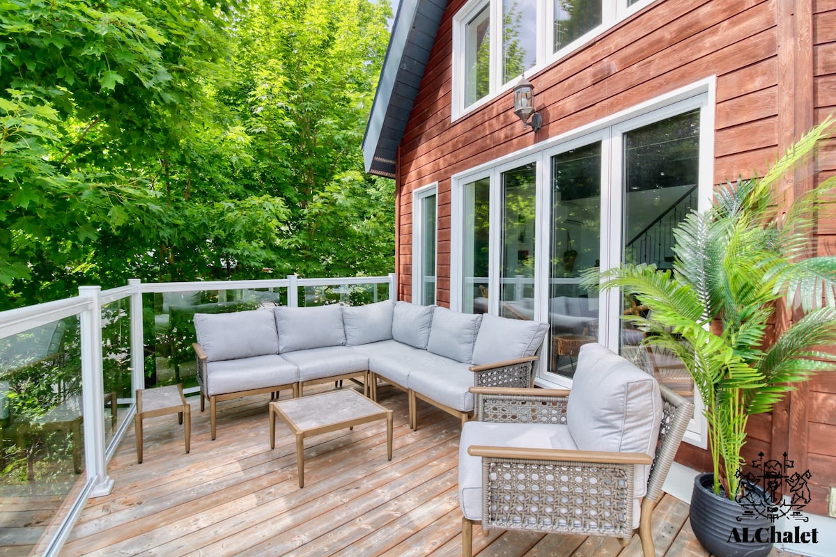 An inviting outdoor seating area is displayed on a wooden deck, featuring a spacious sectional sofa with light grey cushions and a matching coffee table. Lush greenery surrounds the space, complemented by large glass windows of the chalet, allowing for a serene and tranquil atmosphere.