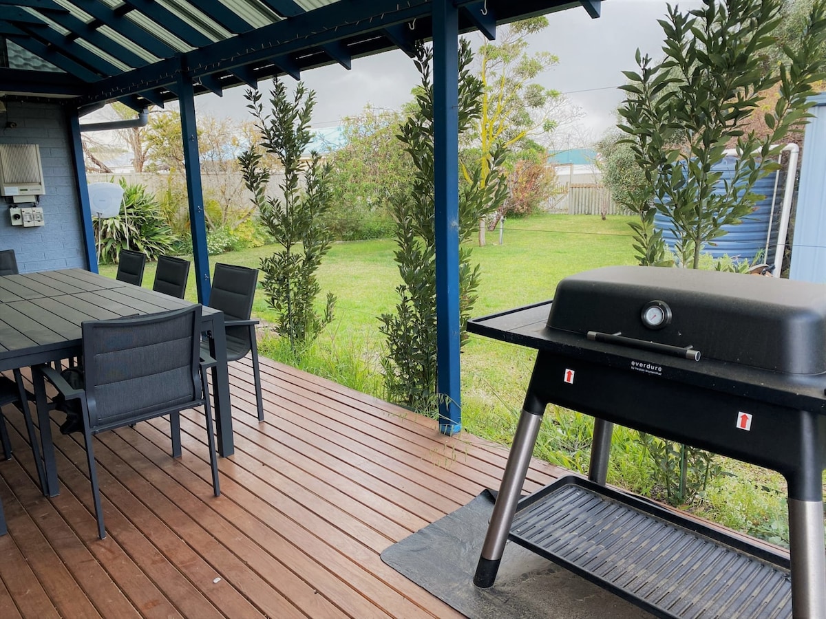 A covered outdoor decking area features a spacious table surrounded by black chairs. A barbecue grill is positioned nearby, with a view of a green lawn and shrubs in the background, creating a comfortable space for outdoor dining and relaxation.
