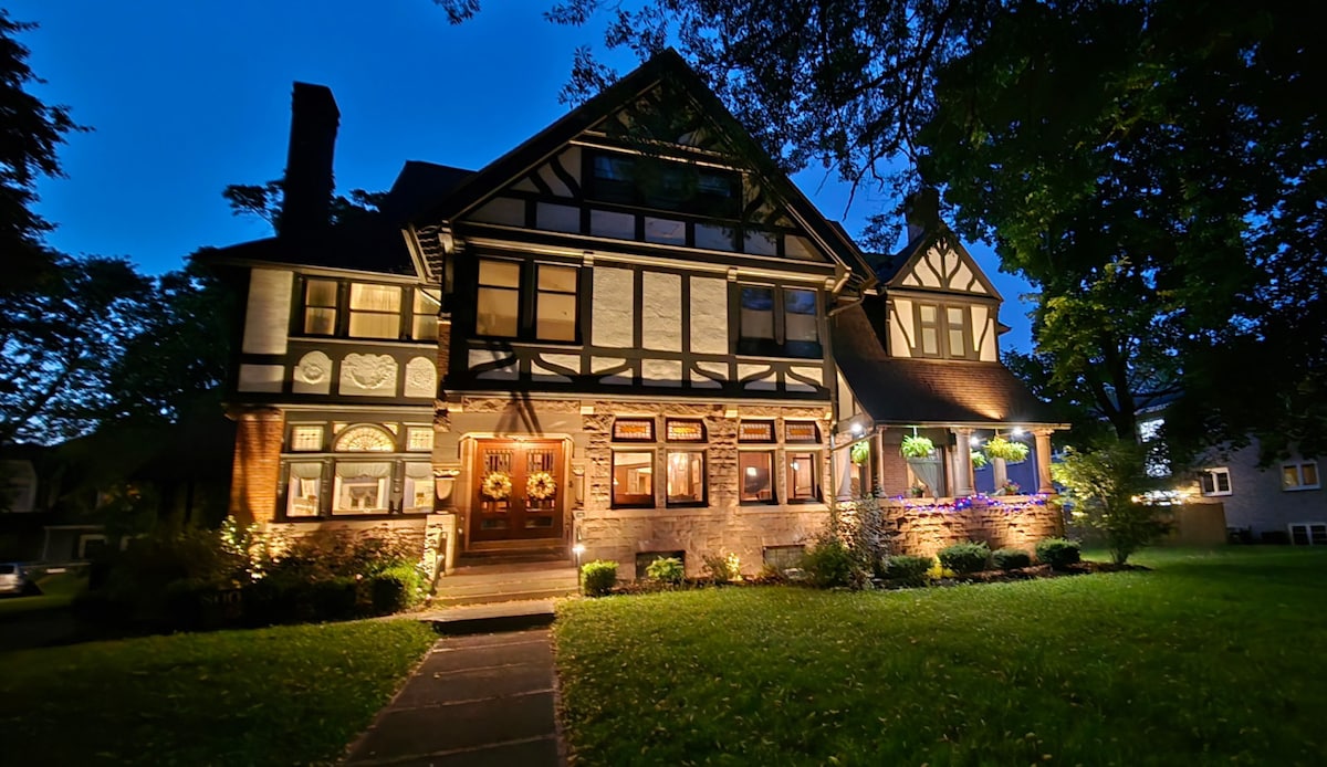 The stately mansion is illuminated at dusk, highlighting its classic architectural details and intricate woodwork. Lush greenery surrounds the house, while light shines from the windows, creating a welcoming ambiance. The inviting entrance features an array of outdoor lighting and seasonal decorations.