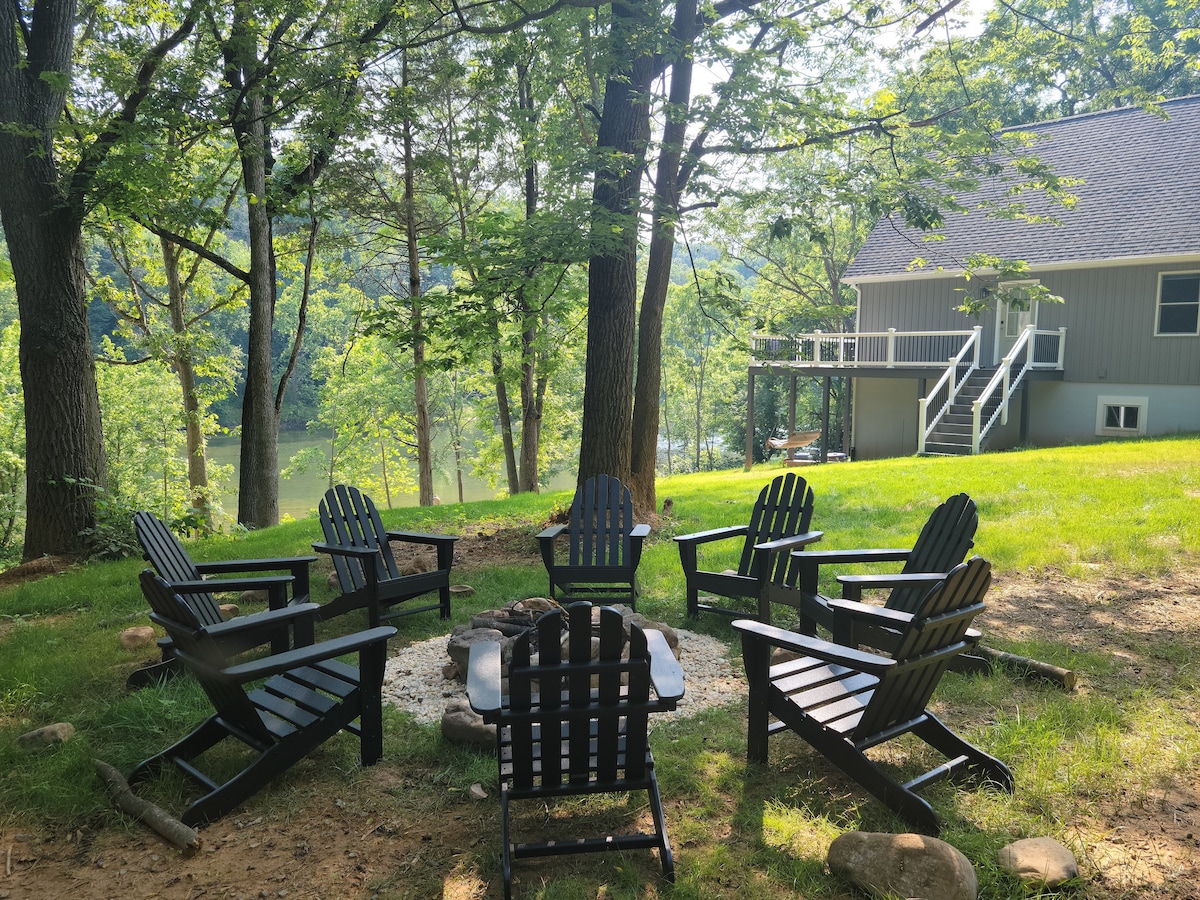 A gathering area is set with six sturdy black Adirondack chairs arranged around a circular fire pit. Lush green grass surrounds the space, while trees provide shade in the background. The river is visible through the foliage, enhancing the serene outdoor setting.