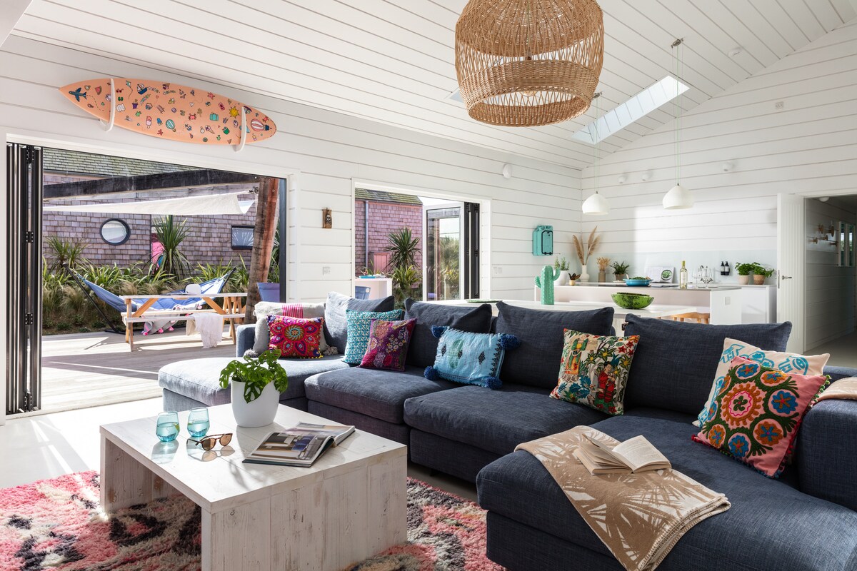 A spacious living area features a deep sectional sofa adorned with colorful throw pillows. Natural light floods the room through large folding doors, revealing a vibrant outdoor space. A rustic coffee table holds a book, with a plant adding a touch of greenery.
