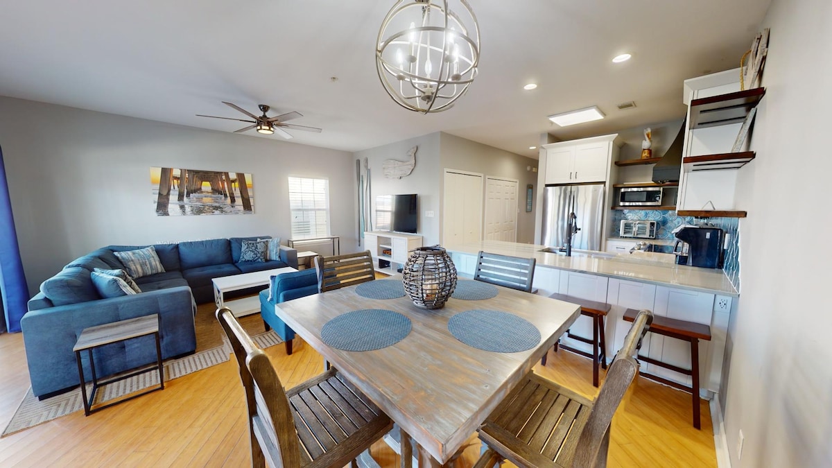 A spacious living and dining area is highlighted by a large blue sectional sofa and a wooden dining table surrounded by sturdy chairs. A decorative light fixture hangs above the table, while an adjacent kitchen features modern appliances and ample storage.