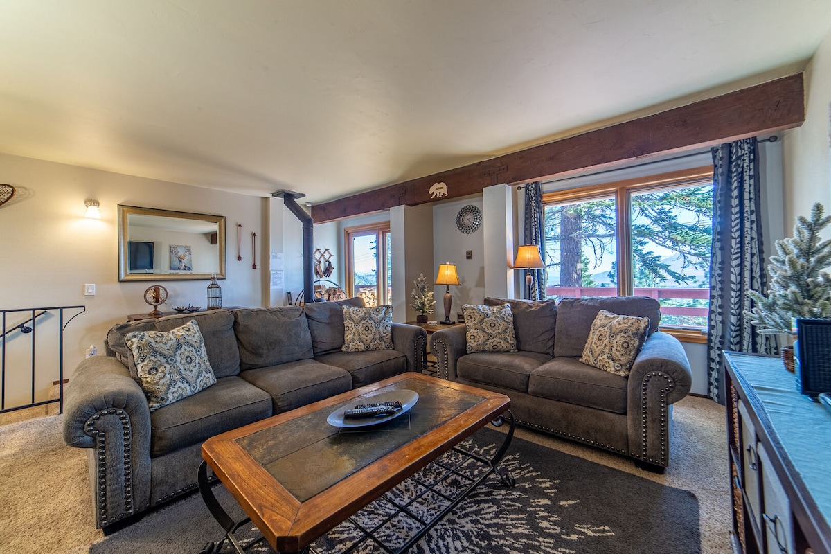 A spacious living area is furnished with comfortable couches and a wooden coffee table. Large windows provide a view of the surrounding trees, and soft lighting from table lamps creates a cozy ambiance. Decorative elements add character to the inviting space.