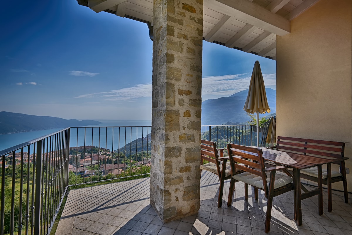 An outdoor terrace is depicted with a wooden table and chairs, accompanied by a large umbrella. The space is overlooking stunning views of the mountains and lake, framed by a stone column and a railing.