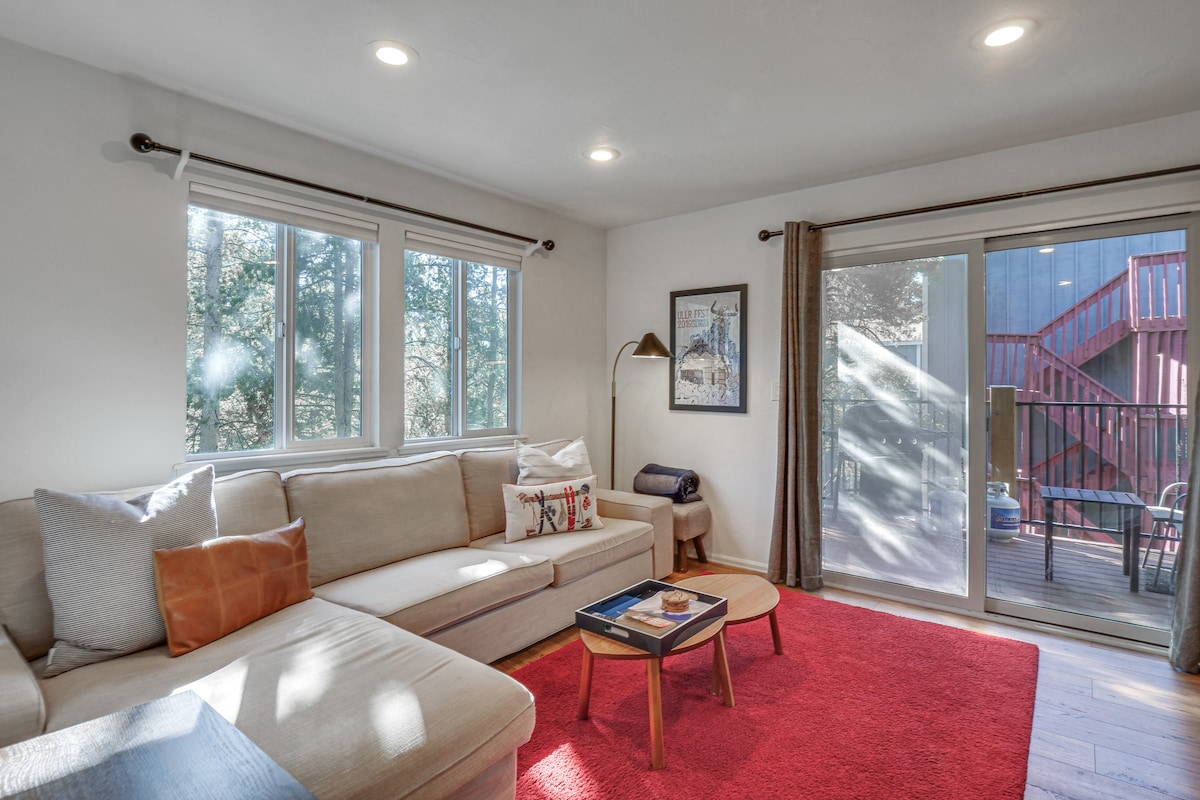 A cozy living area is highlighted by a light-colored sectional sofa, complemented by soft throw pillows. Natural light enters through multiple windows, illuminating the space. A red area rug adds warmth, while a coffee table features books and refreshments. A sliding glass door leads to an outdoor balcony.