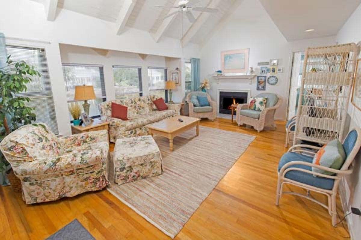 A spacious living area features light wooden floors and large windows that provide natural light. Floral-patterned sofas and armchairs are arranged around a central coffee table. A fireplace is positioned on the right side, complemented by decorative artwork and plants throughout the room.