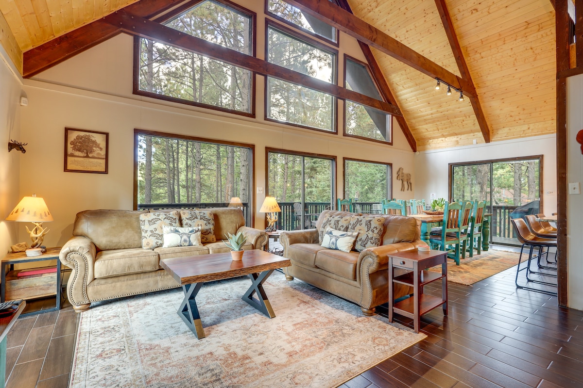 An open-concept living area features high wooden ceilings and large windows, allowing natural light to brighten the space. Two comfortable sofas are positioned around a central table, creating a cozy atmosphere. A view of the outdoors is visible through the surrounding trees.