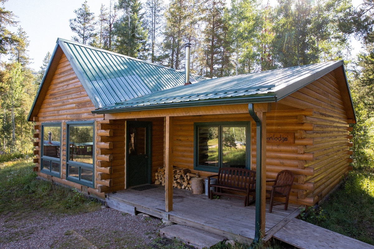 A rustic log cabin features a green metal roof and large windows, allowing for ample natural light. A wooden porch with seating is visible, complemented by a stack of firewood nearby. Surrounding trees provide a natural backdrop.