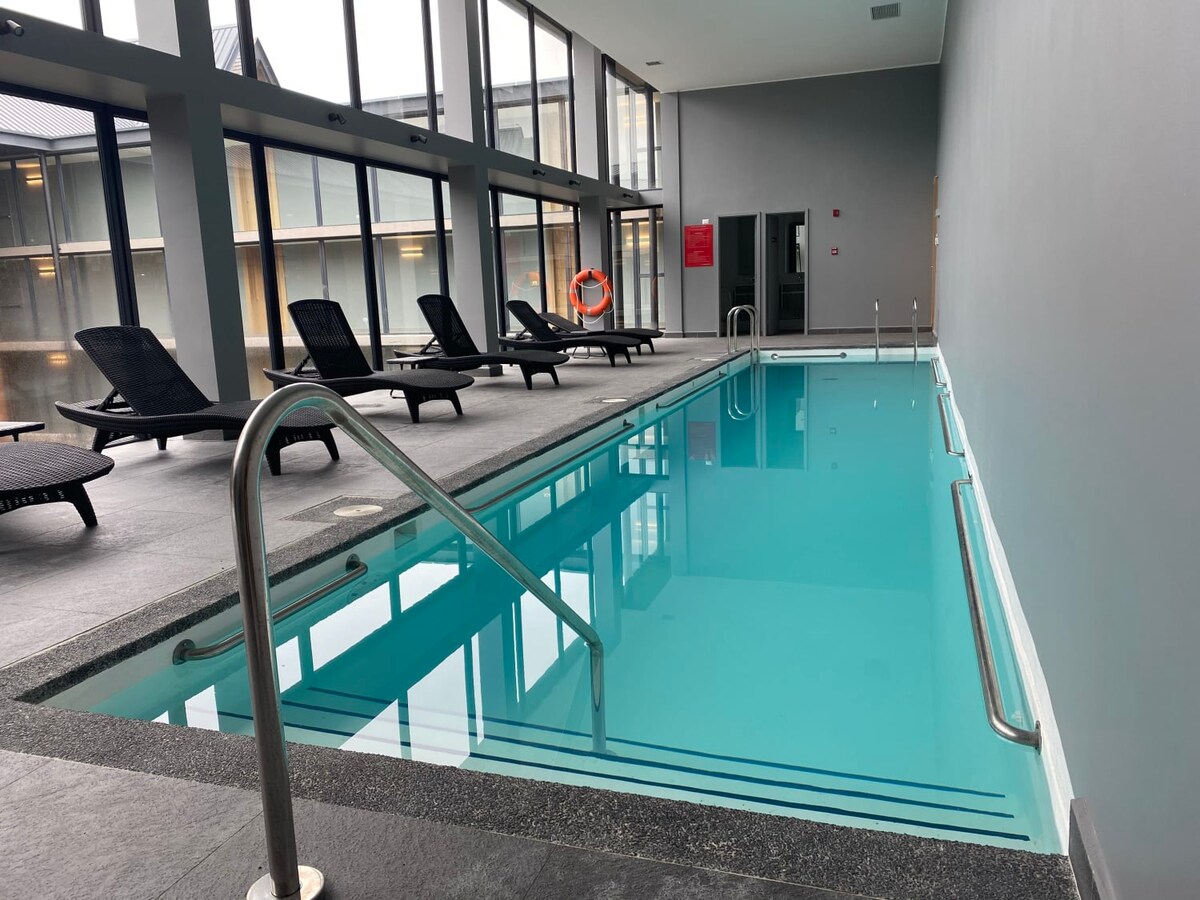 An indoor heated pool is featured, surrounded by several lounge chairs. The pool’s clear water contrasts against the gray walls and flooring. Large windows offer natural light, creating a bright and inviting atmosphere for relaxation.