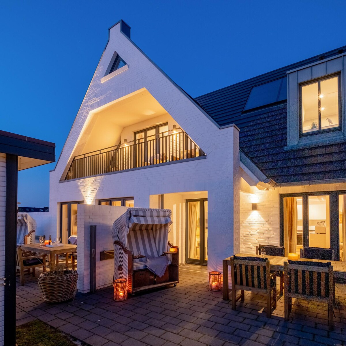 The exterior of a stylish two-story house features a distinctive peak roof and large windows, allowing for ample natural light. A cozy terrace area is furnished with seating and elegant lighting, creating a welcoming atmosphere for outdoor relaxation. The scene is set against a twilight sky.