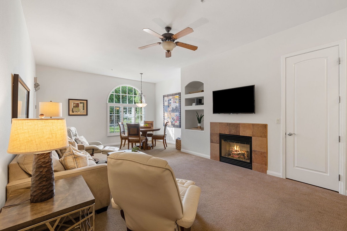 A spacious living area features a comfortable seating arrangement, including a recliner and sofa. A fireplace is set within a stone surround, while a flat-screen TV is mounted on the wall. Large windows allow natural light to fill the space, highlighting a dining area nearby.