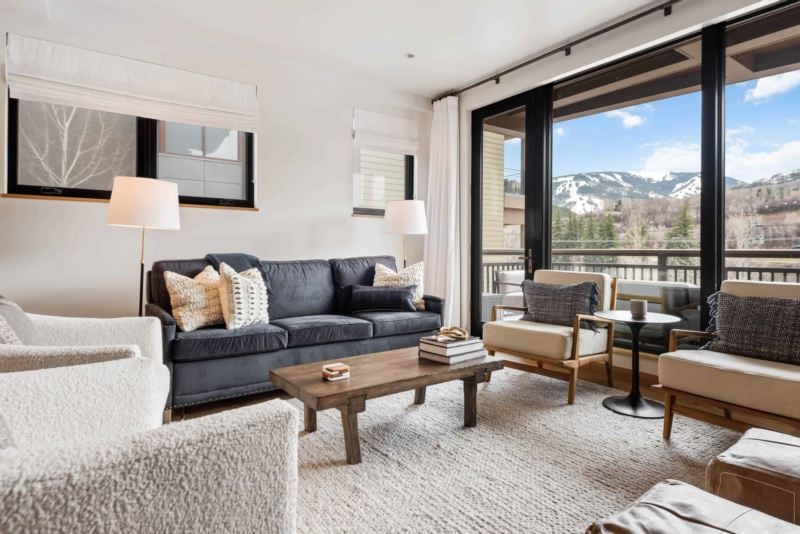 A well-furnished living area features a comfortable dark sofa and two armchairs, oriented around a rustic wooden coffee table. A large window with unobstructed mountain views invites natural light, while soft white curtains add a touch of elegance.