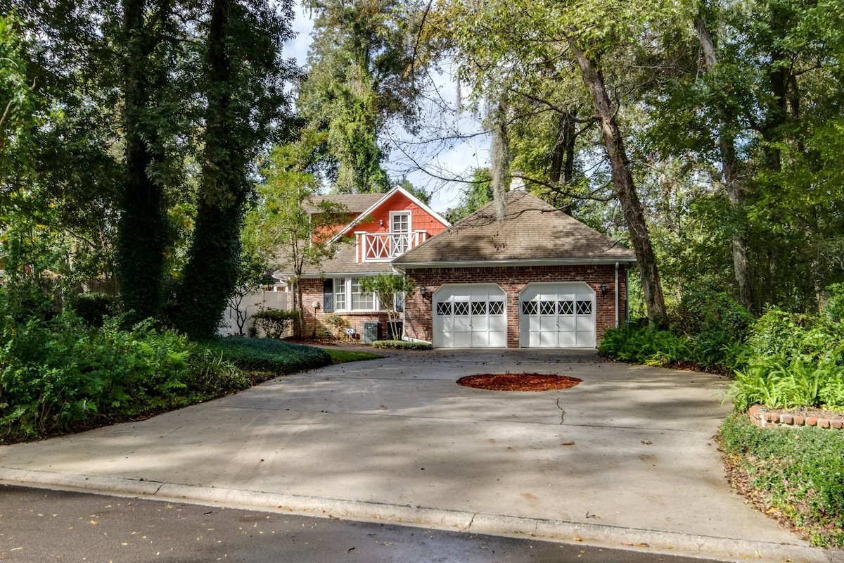 The exterior of the home showcases a charming English cottage style with a red facade and a sloped roof. A large driveway leads to two garages with decorative doors. Lush greenery surrounds the house, highlighted by mature trees and landscaping.