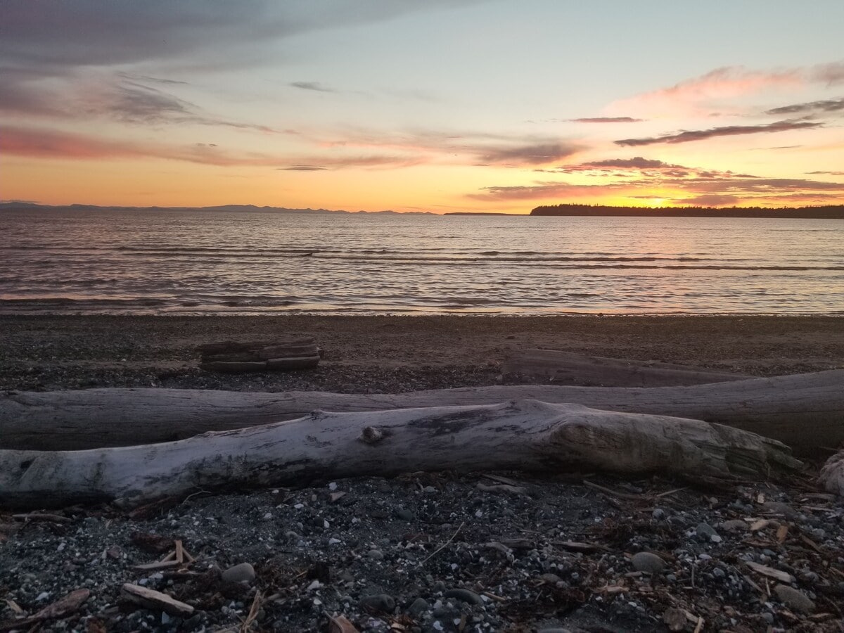 A serene beach scene captures the sunset over calm waters, with the silhouette of distant islands visible on the horizon. Driftwood is scattered along the shoreline, and small pebbles are seen in the foreground, enhancing the natural coastal environment.