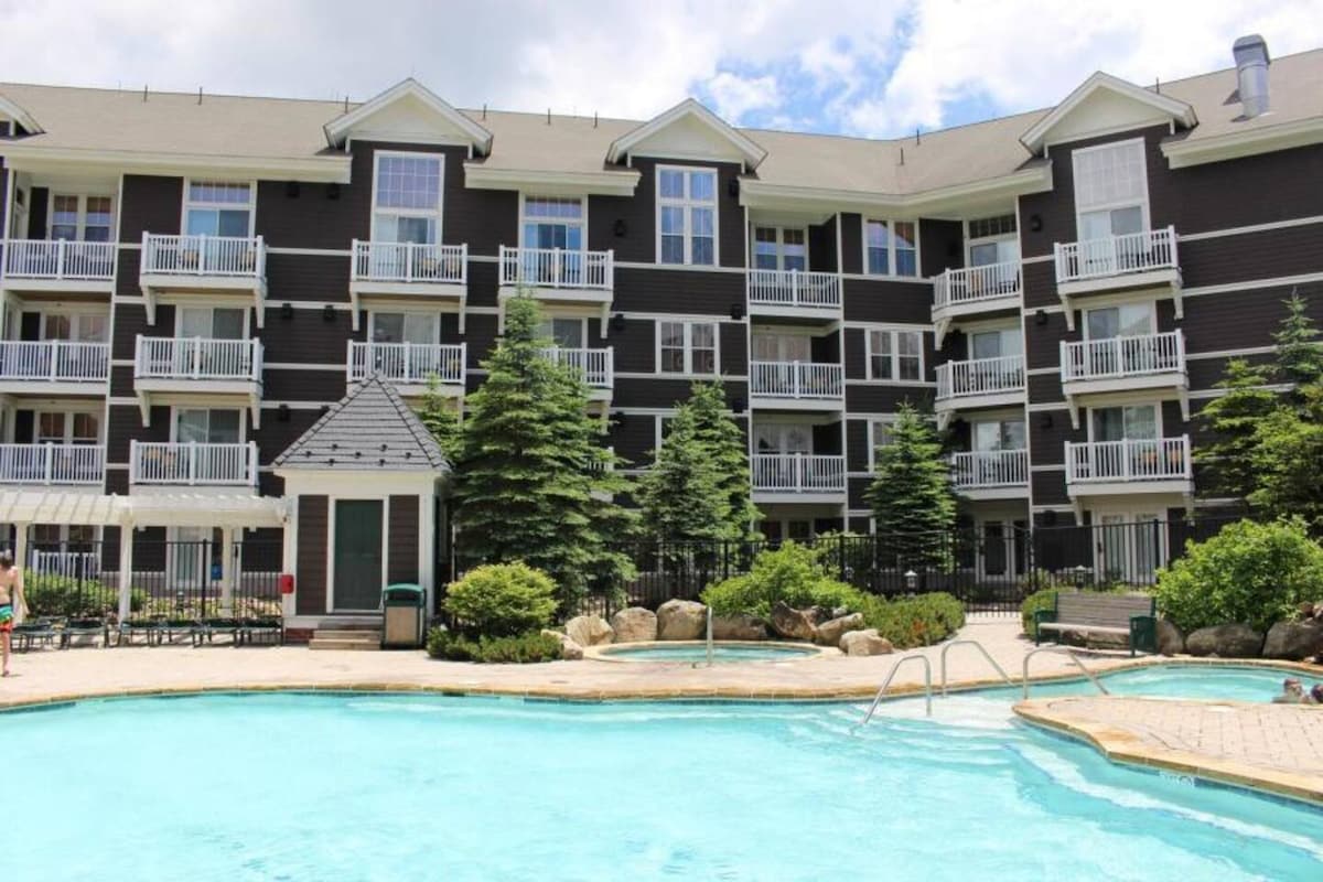 The exterior of the condo building is highlighted, featuring multiple balconies with white railings. A clear swimming pool is positioned in the foreground, surrounded by landscaped greenery and stone accents, providing a serene recreational area.